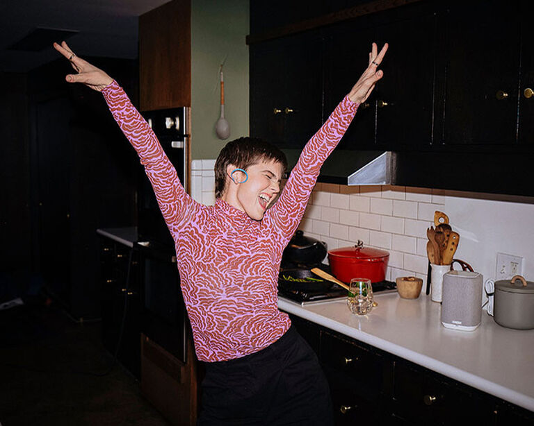 Woman in a patterned pink shirt dancing in the kitchen next to a Denon Home 150